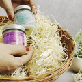 Two jars of eco friendly candles held by a hand over a basket filled with shredded paper.