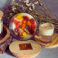 ecofriendly Candle with colorful candy pieces inside, surrounded by dried flowers and another candle on a wooden coaster.