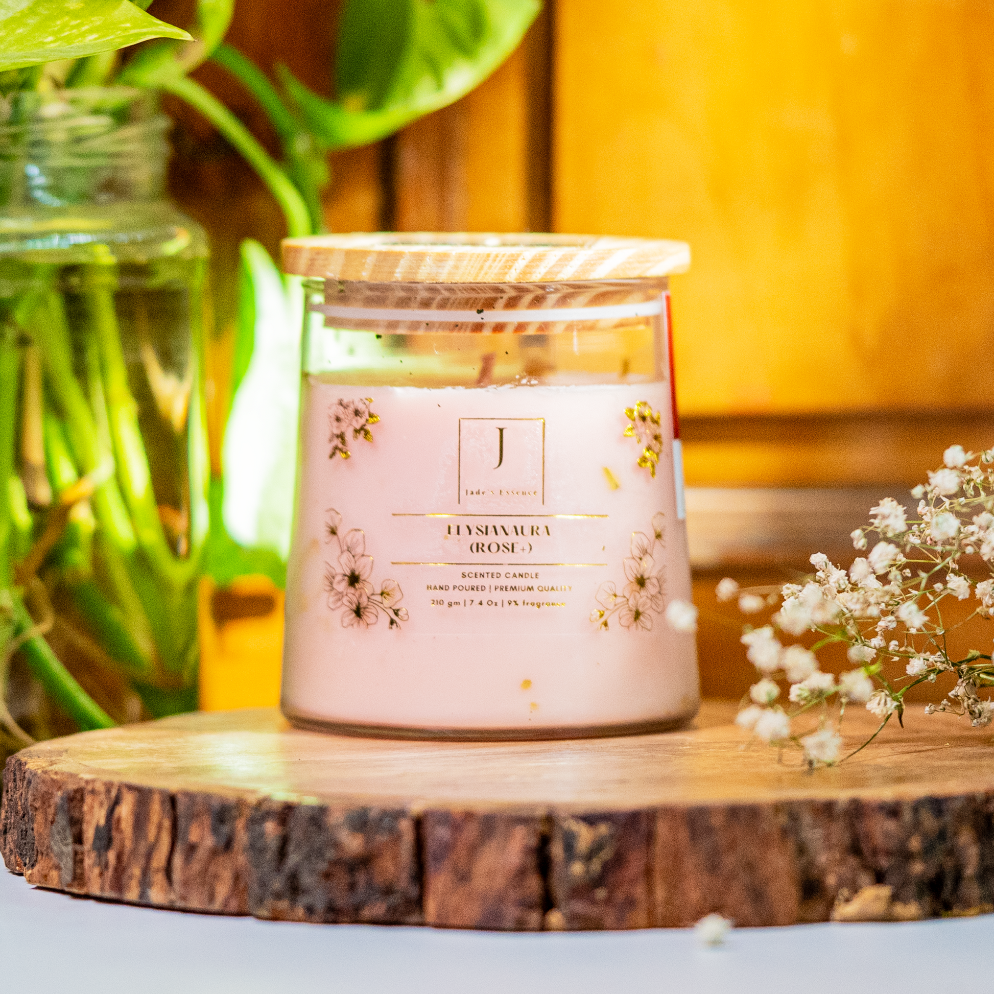 A colorful taper glass Eco friendly candle with gold leaves, placed on a wooden surface, with a blurred background featuring greenery and a wooden object.