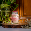An Eco friendly candle candle with gold leaves inside a clear glass jar, placed on a wooden surface, with green foliage and a plant in the background.