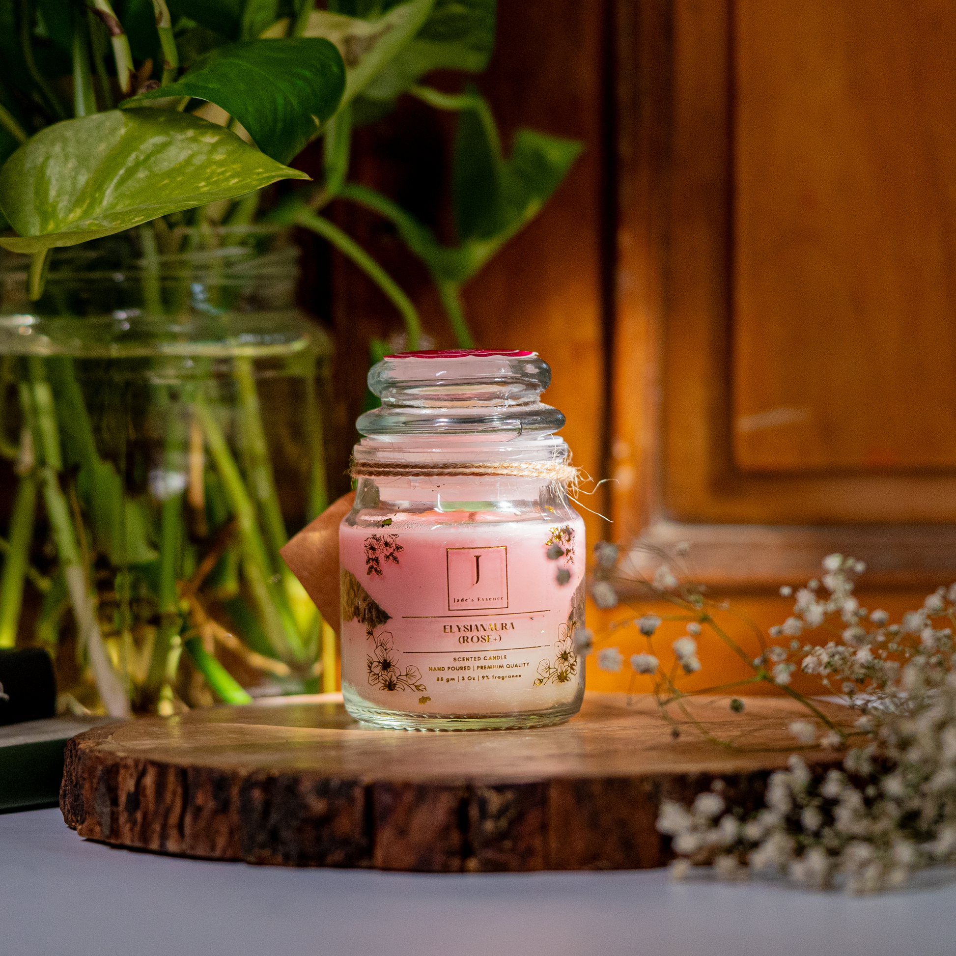 An Eco friendly candle candle with gold leaves inside a clear glass jar, placed on a wooden surface, with green foliage and a plant in the background.