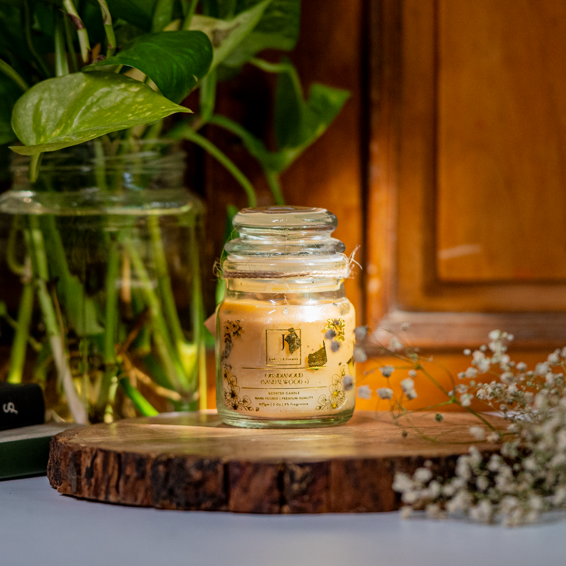 A ecofriendly scented candle with gold leaves inside a clear glass jar, placed on a wooden surface, with green foliage and a plant in the background.