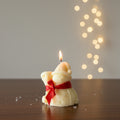 Candle shaped like a bear with a red bow, lit on a wooden surface with a blurred light background.