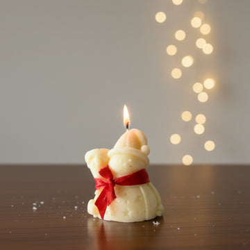 Candle shaped like a bear with a red bow, lit on a wooden surface with a blurred light background.