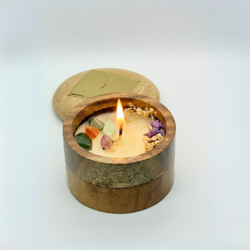 Wooden container with a lit candle inside, featuring small stones and flowers on a light background